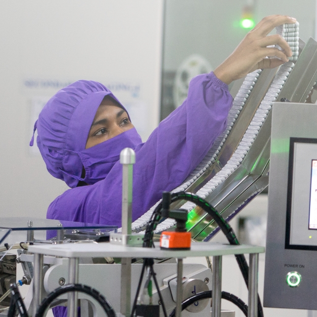Worker in a purple gown, mask, and hairnet handling medicines in a manufacturing facility in Bangladesh.